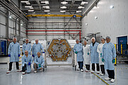 A group of 10 people wearing gloves and hair nets pose for a photo alongside the non-reflective side of a hexagonal mirror segment. Of these, 8 are wearing blue ESO lab coats, and 2 are wearing white lab coats.