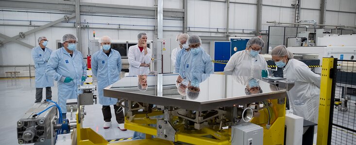 A very shiny hexagonal mirror in the foreground of the image reflects the faces of the people standing around it inside a large, white hall. The people are wearing hairnets and white or blue lab coats; some are wearing face masks and gloves too. The mirror rests on top of a yellow metallic structure, with different electronics coming out of it.